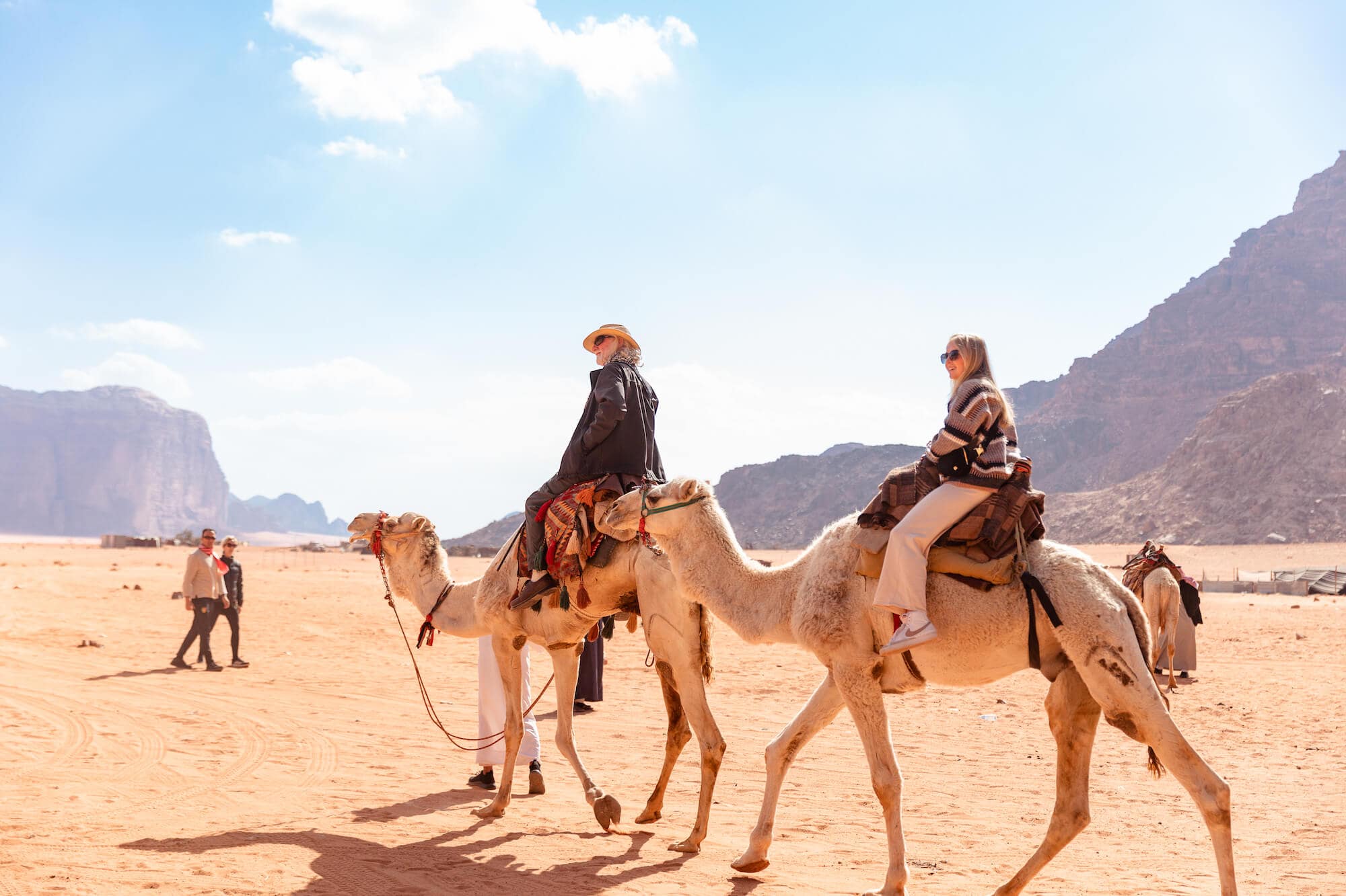 Two people ride camels over desert sand with jagged mountains in the background, while two more people pass by on foot in the middle distance.
