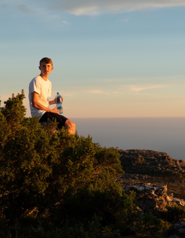 A young man sits on a rocky rise overlooking the ocean at sunset.