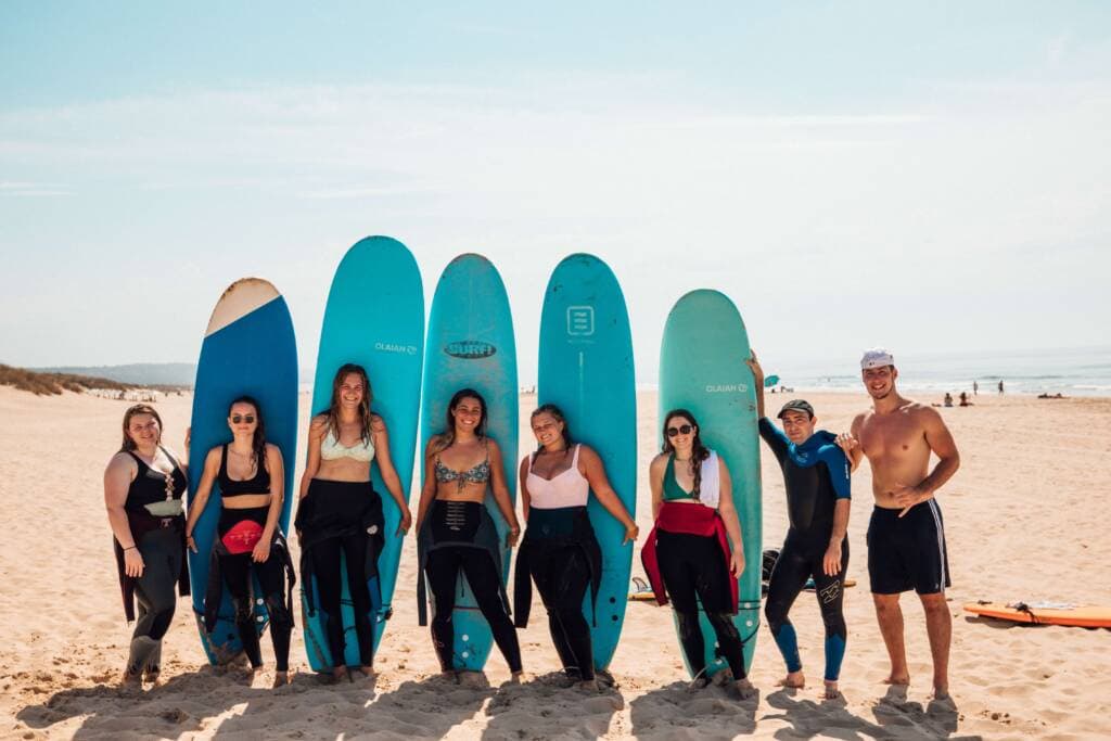 About a half dozen people in swimsuits pose with blue surfboards on a sunny beach.