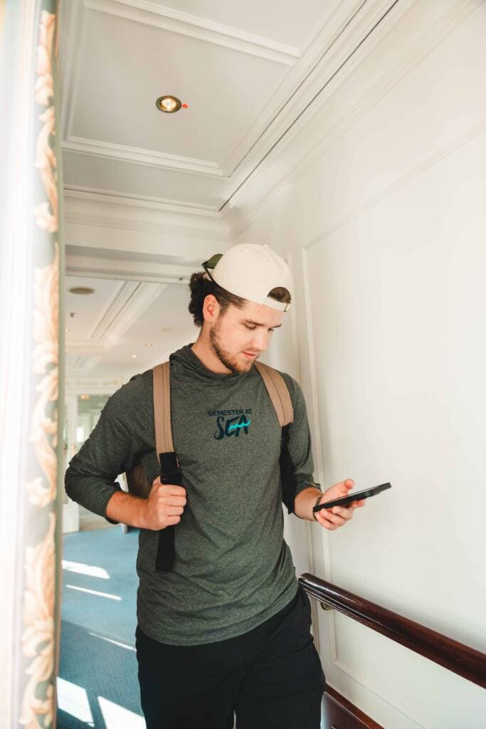 A young man wearing a green shirt, a backwards baseball cap, and a tan backpack looks at a cellphone.