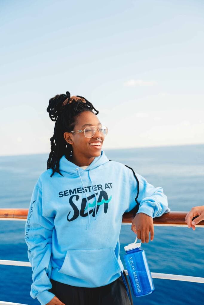 A young woman wearing glasses, a nose ring, and a bright blue hoodie leans against the rail of a ship while holding a blue plastic water bottle. Blue water is visible behind her.