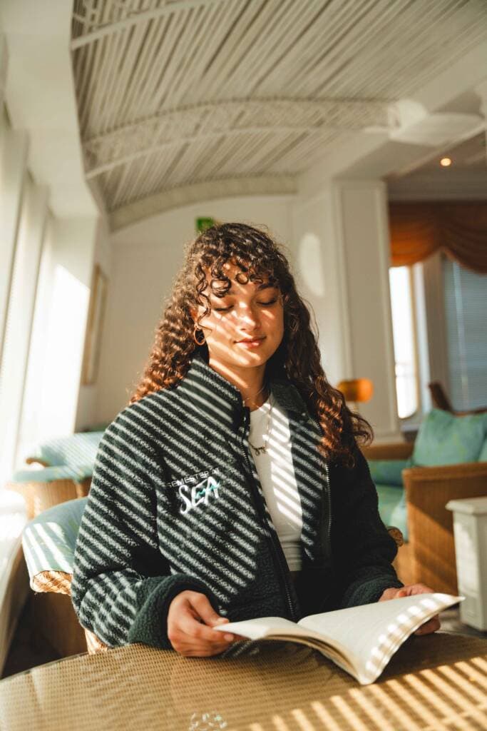 A young woman wearing a green fleece sits next to a sunny window, reading a book. Thin bands of sunlight fall across her.