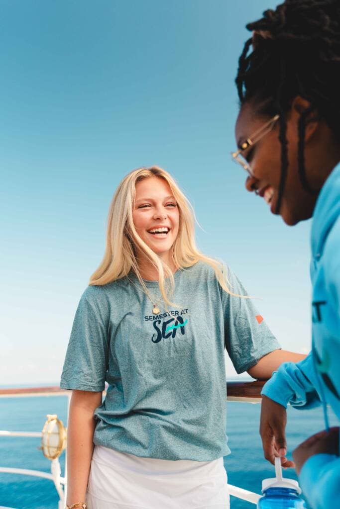 Two young women stand at the rail of a ship, laughing together. One wears a blue-green t-shirt and the other wears a bright blue hoodie.