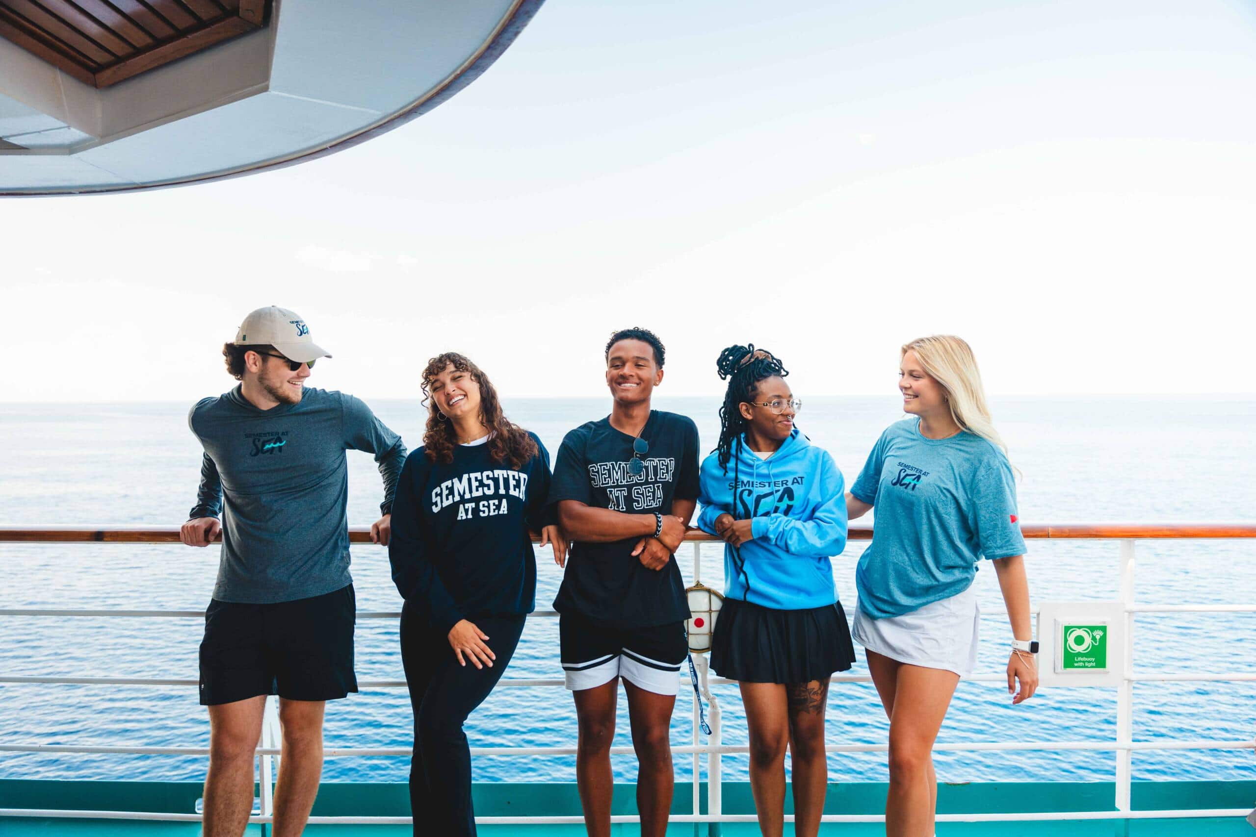 Five young people wearing different shades of blue stand against the railing of a boat, with blue water visible behind them.