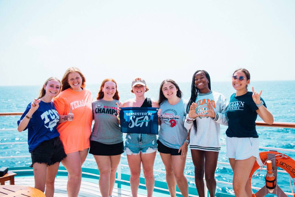 Seven young women standing on a ship's deck with the ocean in the background. They are smiling and holding a 'Semester at Sea' flag Each woman is wearing a different university shirt, representing various schools including Texas, USC, and Miami.