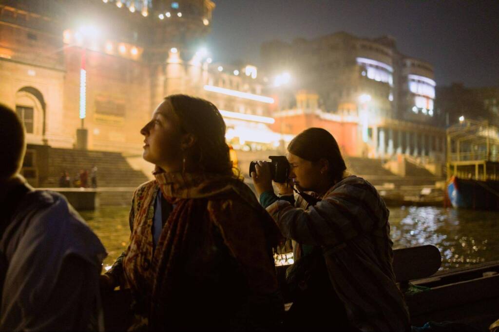 Two young women, one holding a camera, sit in a river boat at nighttime, gazing at the brightly lit buildings onshore. 