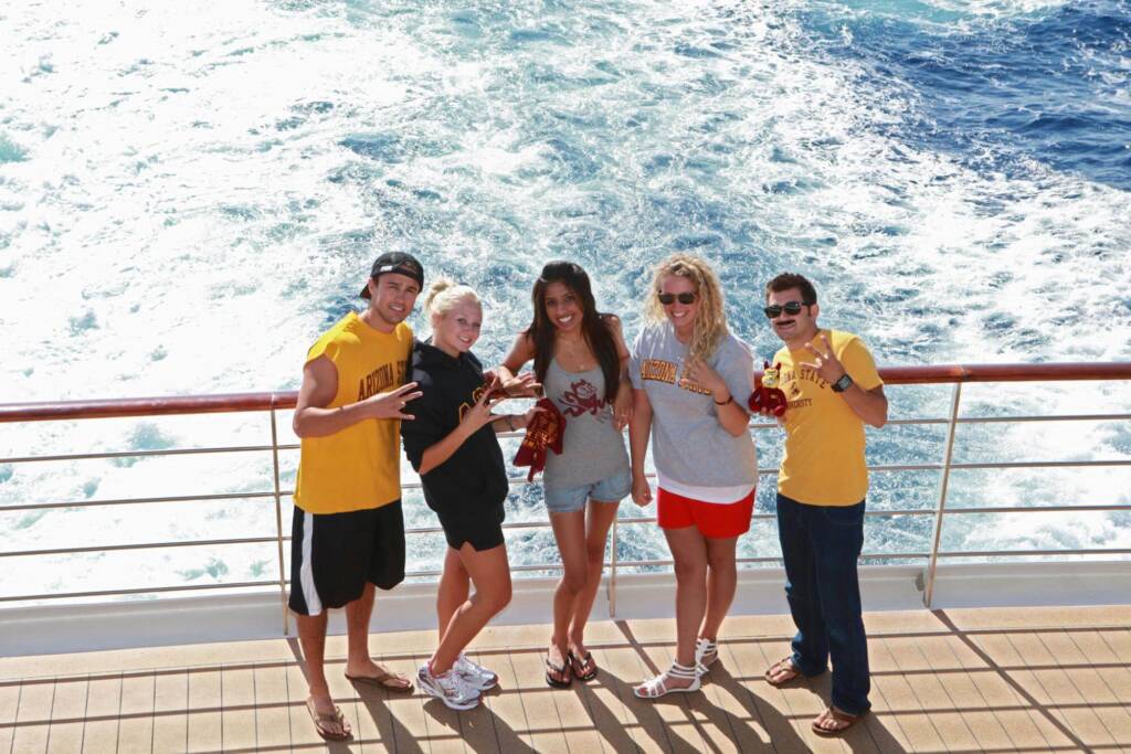 Five ASU study abroad students stand side-by-side, each wearing a different shirt or sweatshirt from their school and flashing the hand sign for the ASU Sun Devils’ sports teams. They are standing on the sunny back deck of a ship, with the ship’s wake visible in the water behind them.