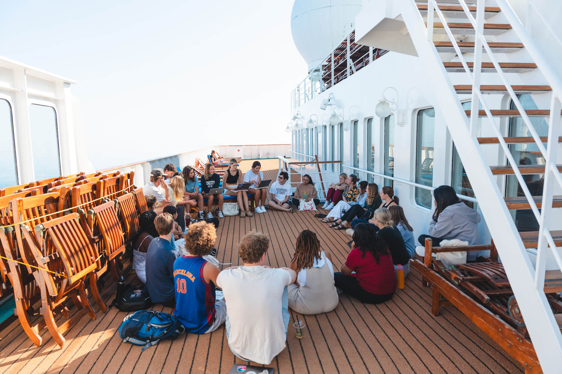 About twenty students sit in a large circle on the deck of a ship, some with laptops propped on their knees. The sea is just visible beyond the ship’s railing. 