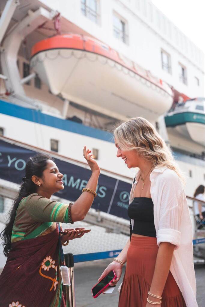 Two women in colorful outfits stand facing each other; one applies a bindi (red dot) to the other’s forehead.