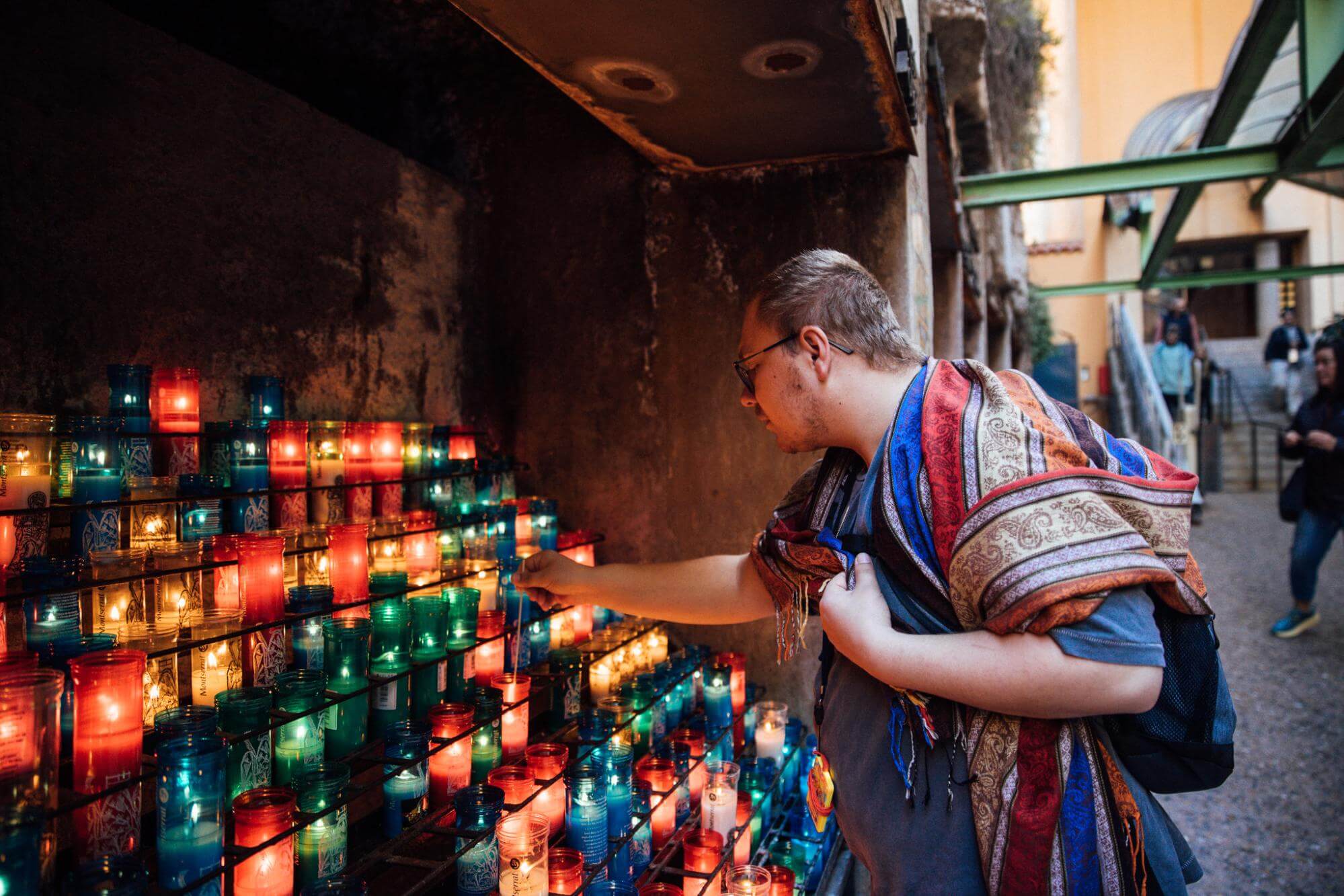 A man wearing a colorful shawl lights a candle in a shrine filled with multicolor glass candle holders.