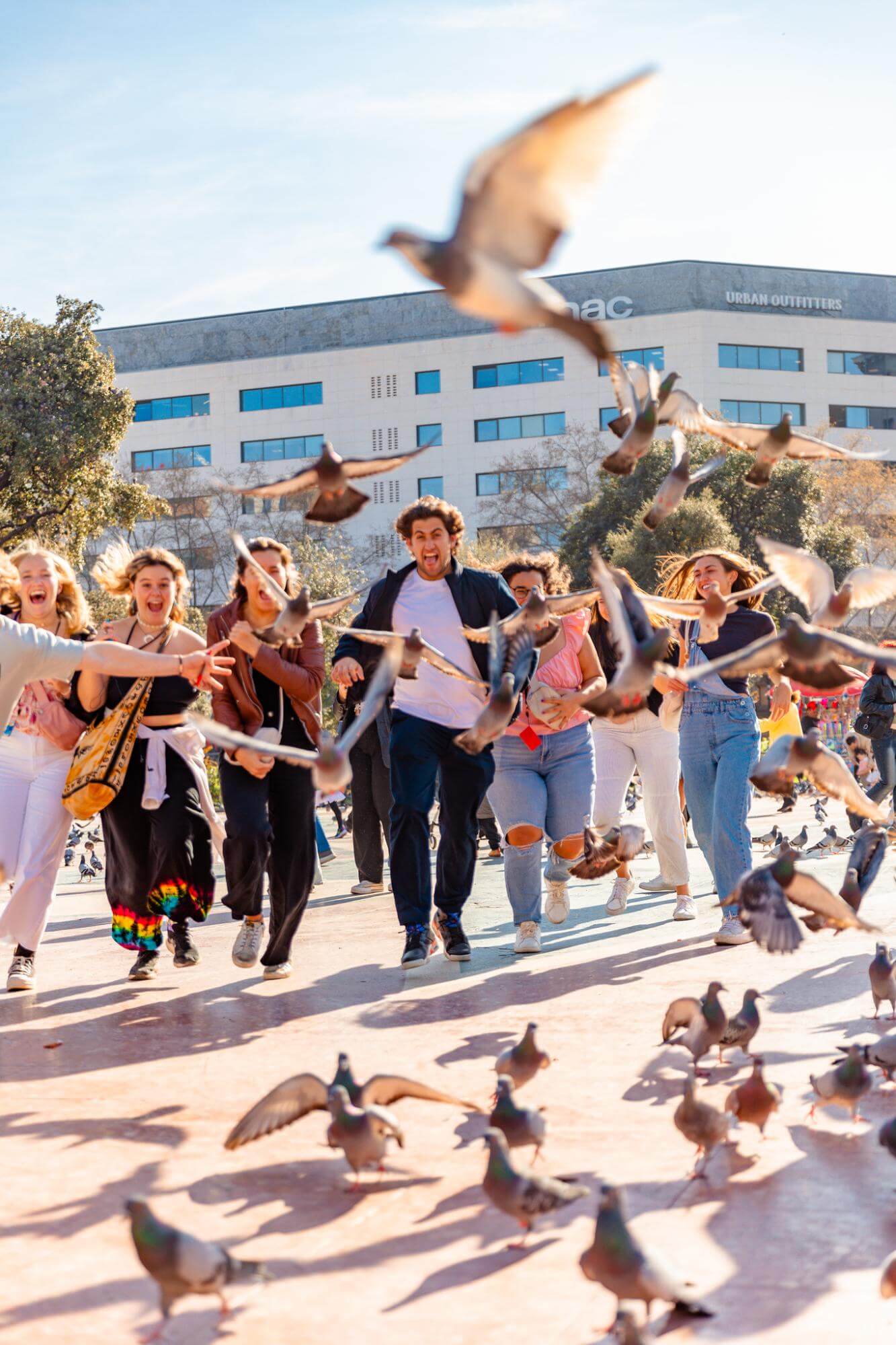 A group of young people run through a city square as a flock of pigeons takes flight.