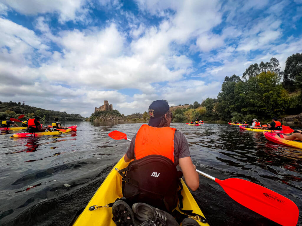 A person in an orange life vest and backwards baseball hat paddles a yellow kayak through a calm body of water, toward a castle perched on the distant shore. Other kayakers are visible on either side.