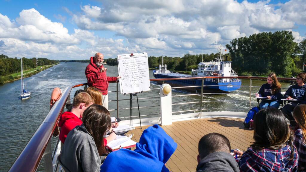 A professor speaks next to a large pad of paper on an easel, surrounded by students on the sunny deck of a ship. They are sailing down a canal as other boats, both large and small, sail past them. 