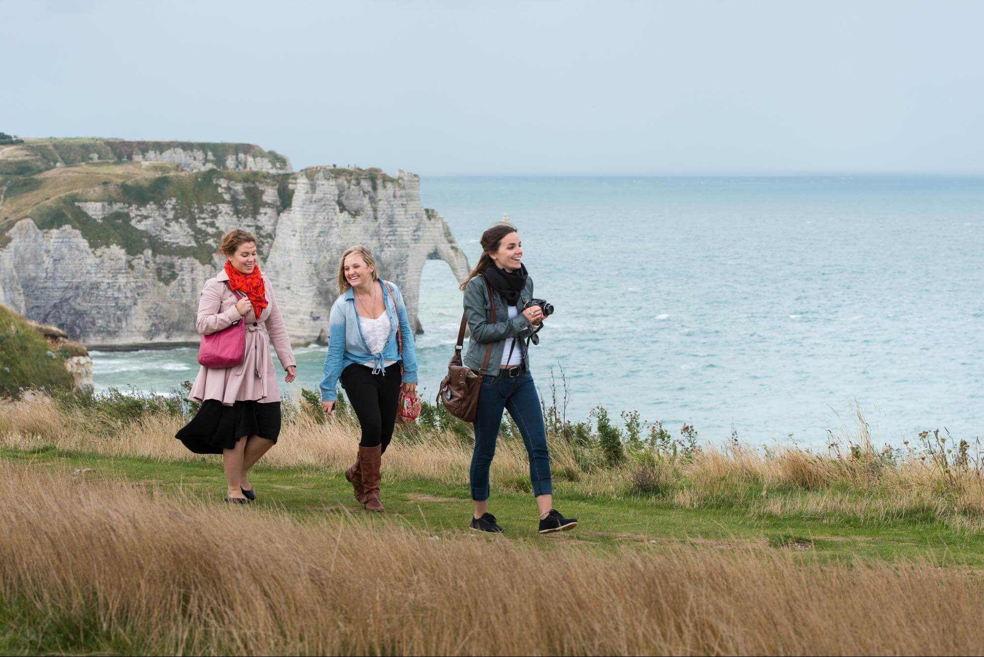 Three young women walk along a grassy path next to the ocean. A cliff face with a striking natural stone bridge looms above the water in the background. 