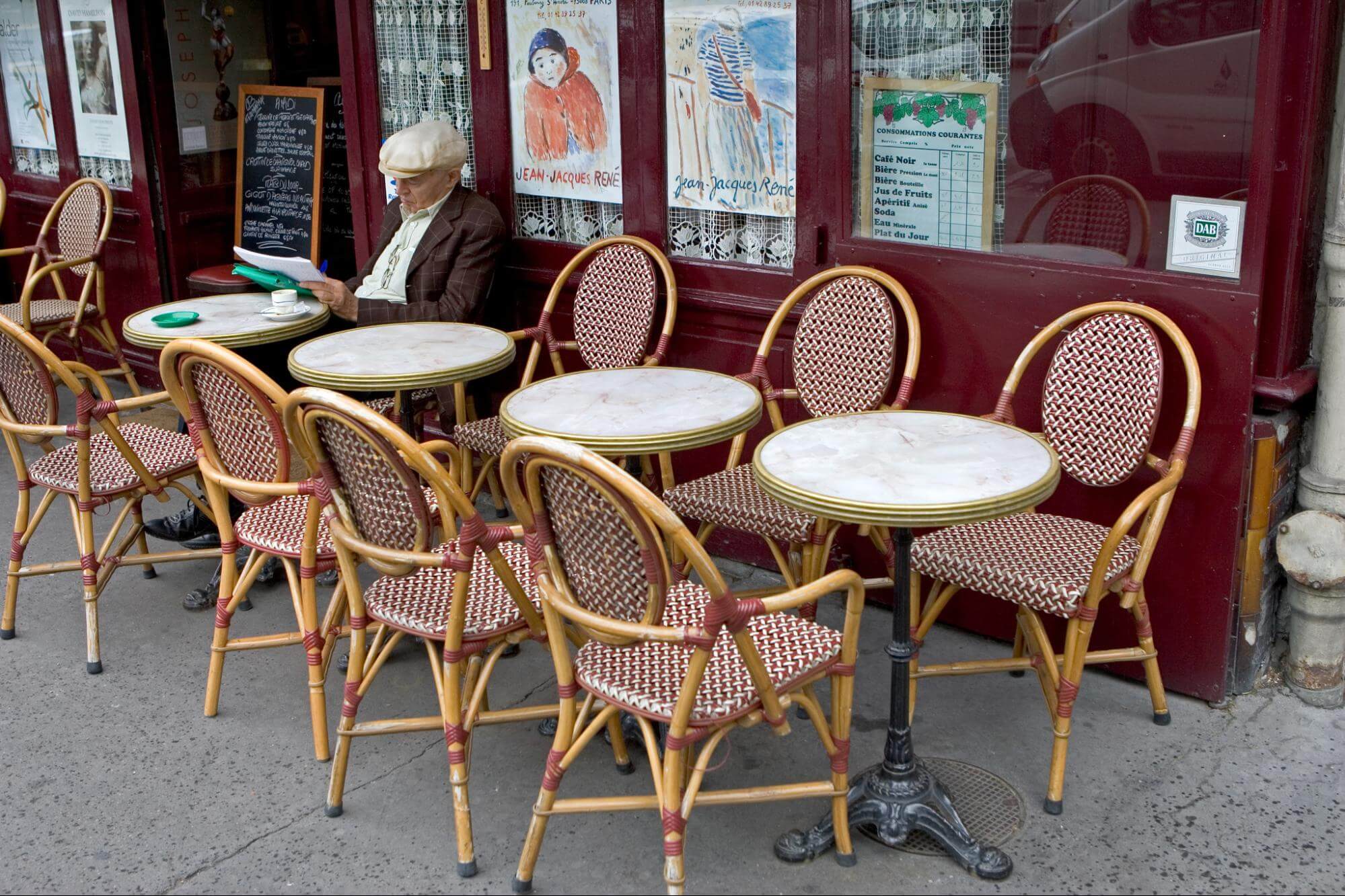 An older gentleman reads a paper outside a cafe. The sidewalk is lined with red and white cane chairs and small, round marble-topped tables. Paintings by Jean-Jacques René are displayed in the cafe windows. 