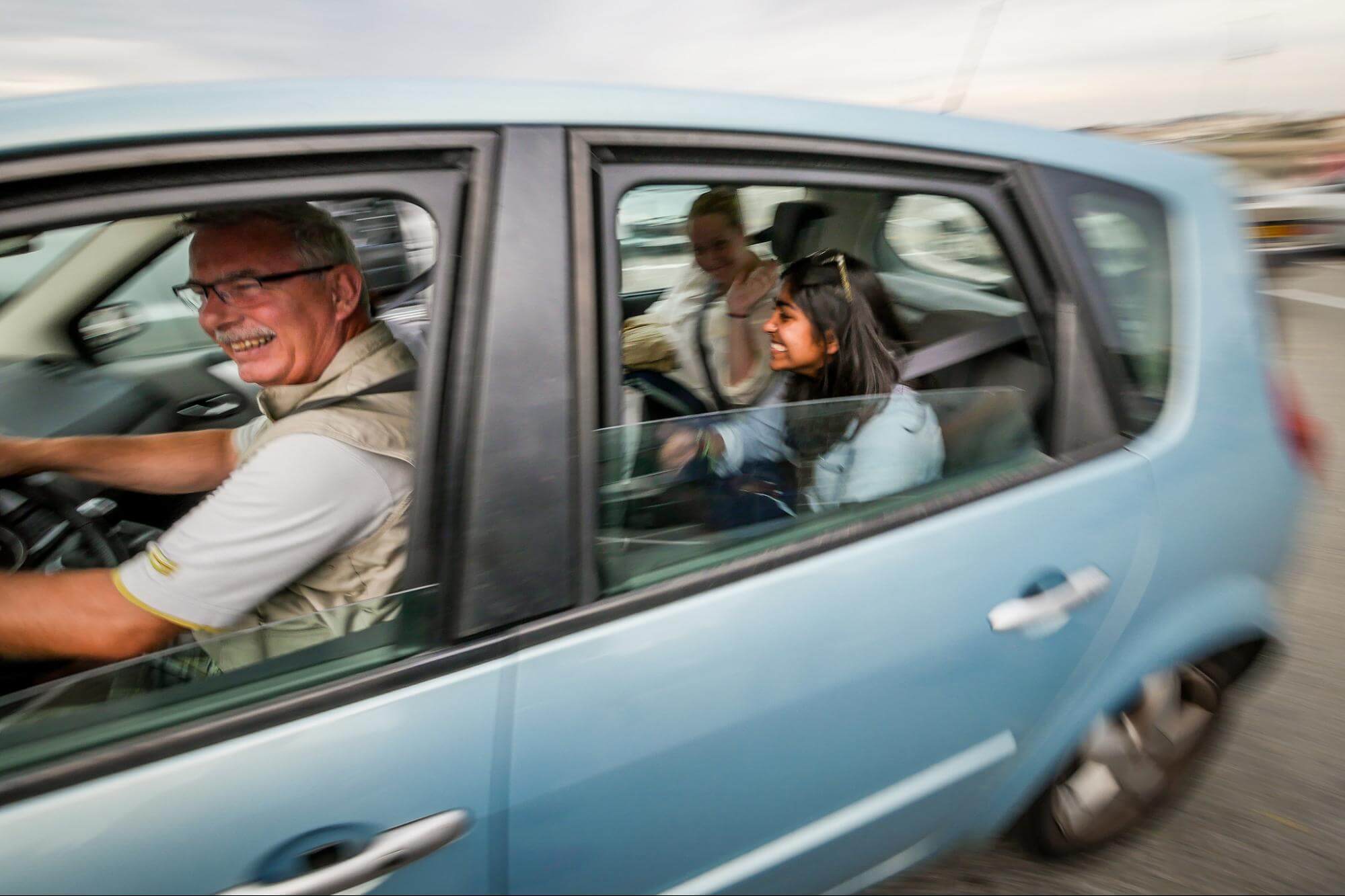Two young women ride in the back of a small blue car driven by an older gentleman. The background of the photo is blurred, indicating the car is in motion. All subjects are smiling.