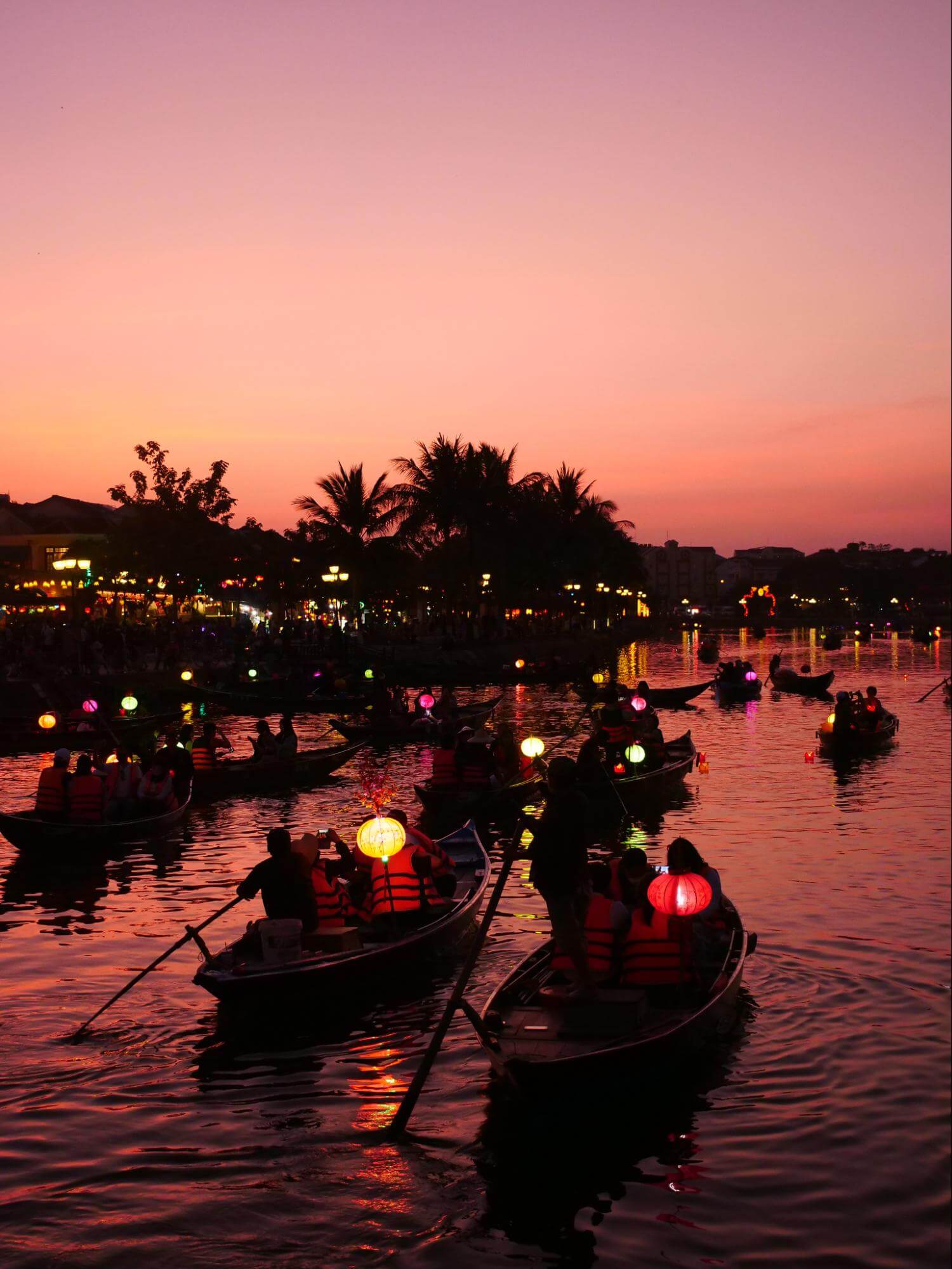 Dozens of small boats float on the water, silhouetted against the sky just after sunset. Each boat carries several passengers and a glowing paper lantern.